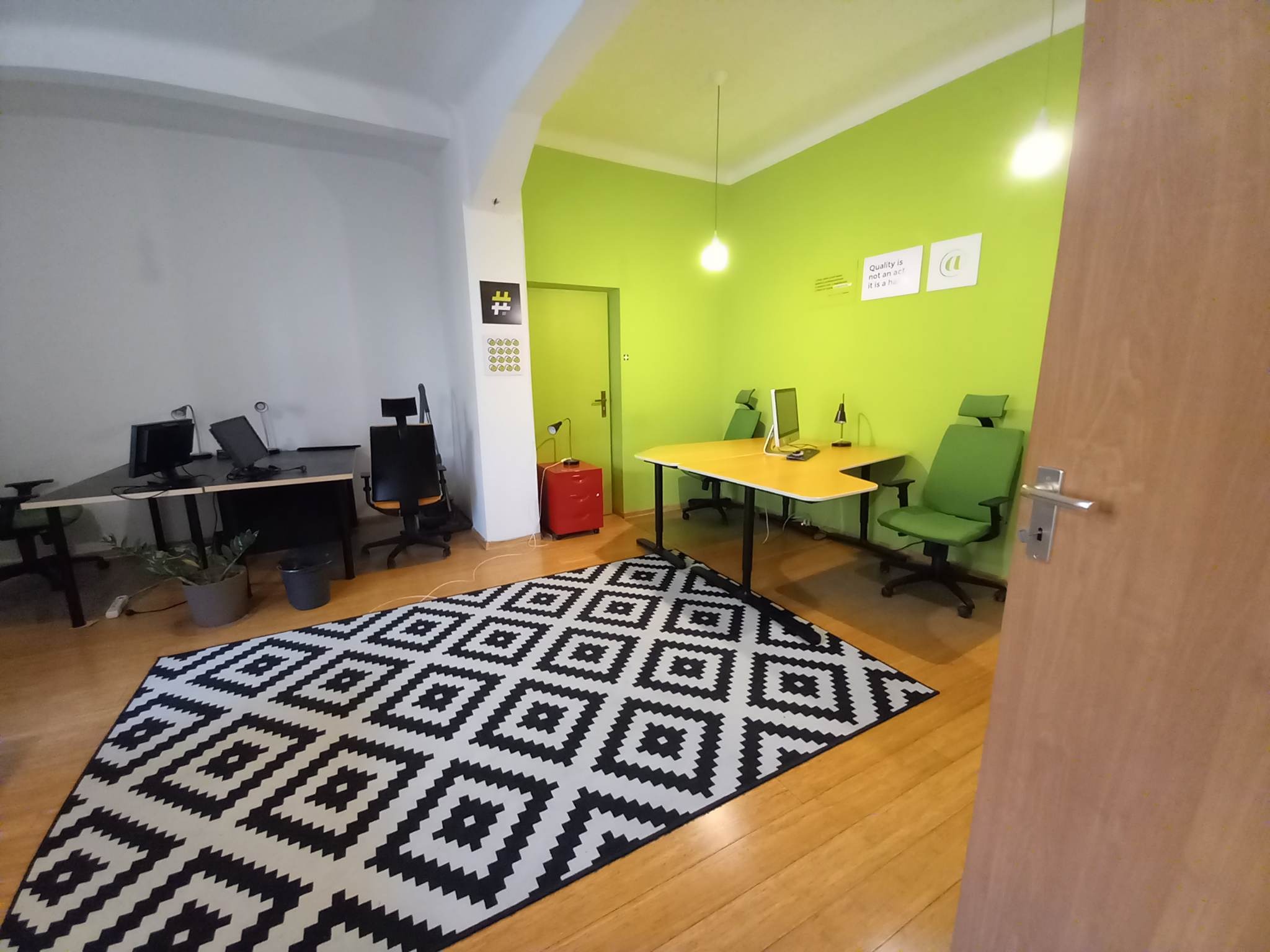 Jasne biuro z zielonymi ścianami, biurkami, krzesłami i czarno-białym geometrycznym dywanem na drewnianej podłodze.
