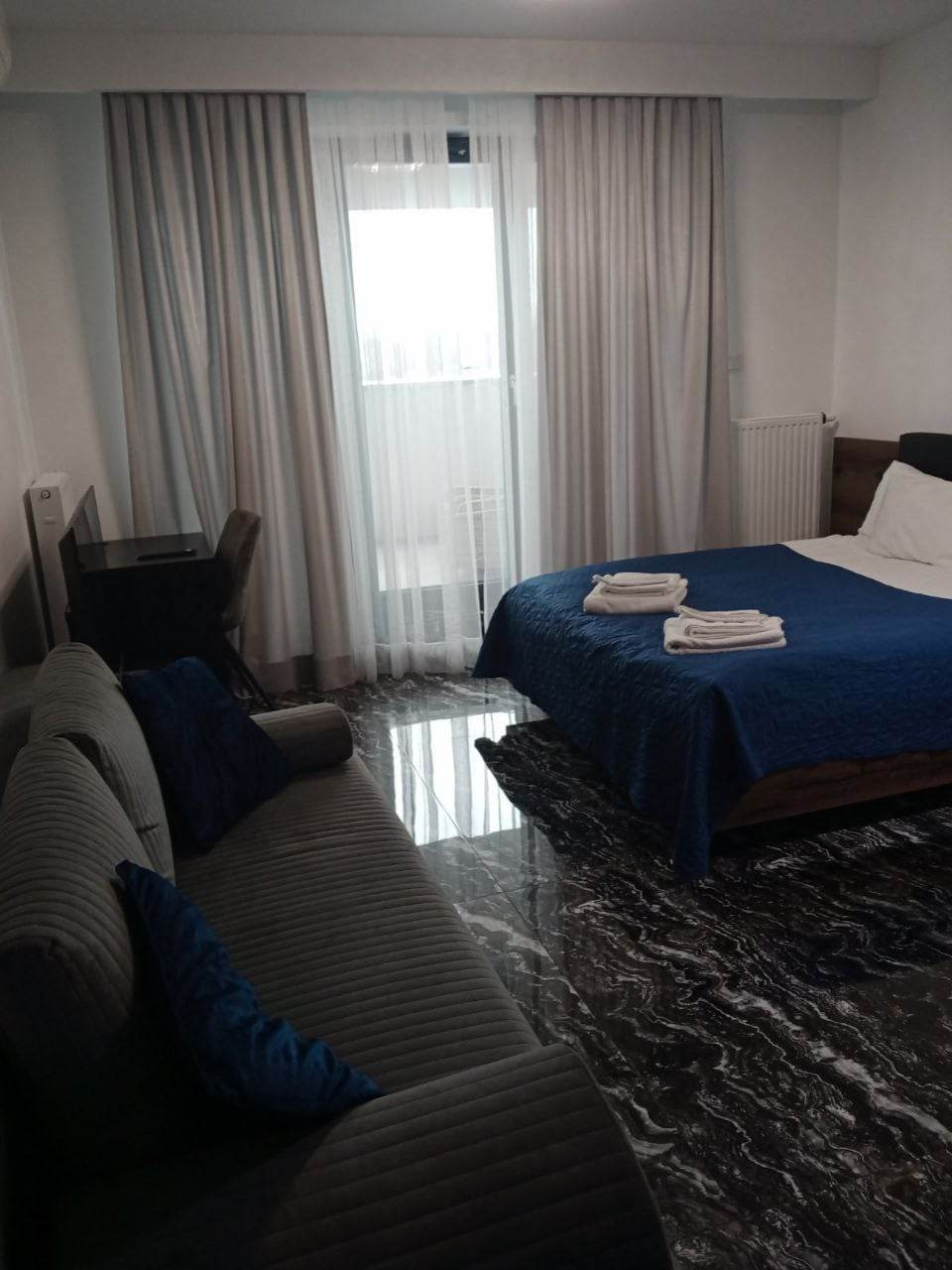 Pokój hotelowy z łóżkiem z niebieską narzutą, szarą sofą, biurkiem i marmurową podłogą odbijającą światło z okna.