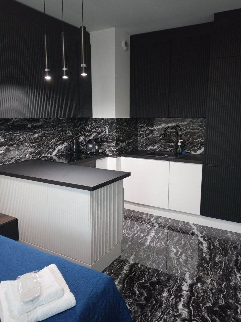 Nowoczesna kuchnia w czerni i bieli z marmurową podłogą, widok na wyspę kuchenną z białymi frontami i czarnym blatem, oraz fragment łóżka z niebieską narzutą i stertą białych ręczników.