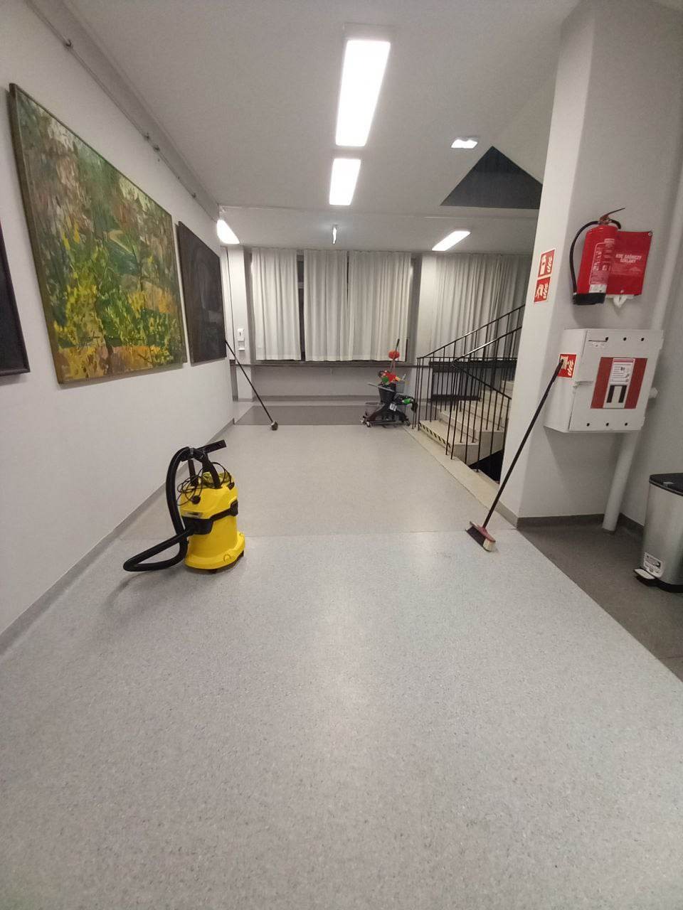 Korytarz biurowy z żółtym odkurzaczem przemysłowym na pierwszym planie, mop oparty o ścianę, w tle schody, obrazy na ścianie i kosz na śmieci.