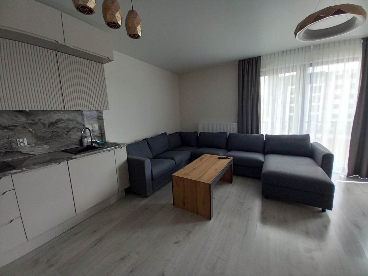 Wnętrze nowoczesnego salonu z szarą narożną sofą, drewnianym stolikiem kawowym z metalowymi nogami, jasnymi panelami podłogowymi i minimalistyczną kuchnią w tle, oświetlonego przez designerskie...