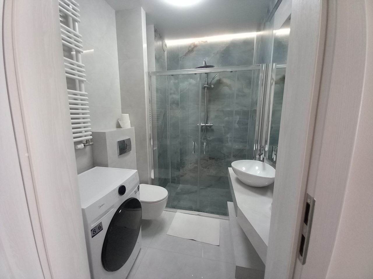 Jasna, nowoczesna łazienka z kabiną prysznicową ze szklanymi drzwiami, umywalką nablatową, pralką i grzejnikiem drabinkowym.