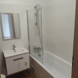 Wykończona łazienka z białą wanną z parawanem nawannowym, umywalką z szafką i lustrem w folii ochronnej, wyłożona białymi płytkami.