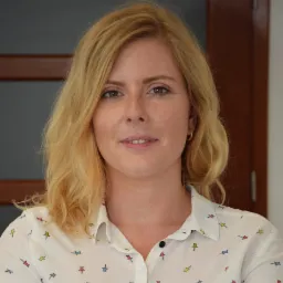 Portret kobiety o jasnych włosach, ubranej w białą koszulę w kolorowe pinezki, z delikatnym uśmiechem i złotymi kolczykami kołami.