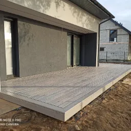 Nowoczesny taras z desek kompozytowych w odcieniach szarości, przylegający do domu w trakcie wykończenia, z widocznymi przewodami na elewacji i niezagospodarowanym terenem wokół.