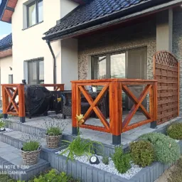 Nowoczesna drewniana balustrada tarasowa z ozdobnymi panelami w Czarna Białostocka, otoczona zielenią i kwiatami, z widocznym fragmentem domu w tle.