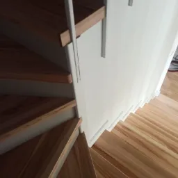 Kątowe ujęcie drewnianych schodów spiralnych z jasnego drewna z białymi wspornikami i ścianą, widok z góry na dół.