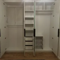 Wnętrze białej szafy wnękowej z otwartymi drzwiami, widoczne półki, szuflady z jasnymi frontami, metalowe kosze na akcesoria oraz drążek na ubrania, podłoga z paneli.