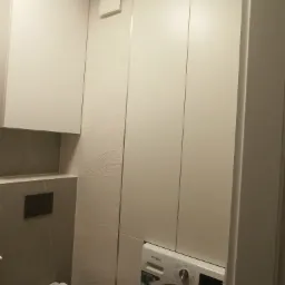 Biała zabudowa meblowa w łazience z pralką Whirlpool wkomponowaną w szafę, obok toaleta i płytki z delikatnym wzorem fal.