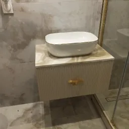 Biała, owalna umywalka nablatowa z delikatnym złotym obramowaniem, osadzona na beżowej szafce łazienkowej z ryflowanym frontem i złotym uchwytem w kształcie liścia, na tle marmurowych płytek...