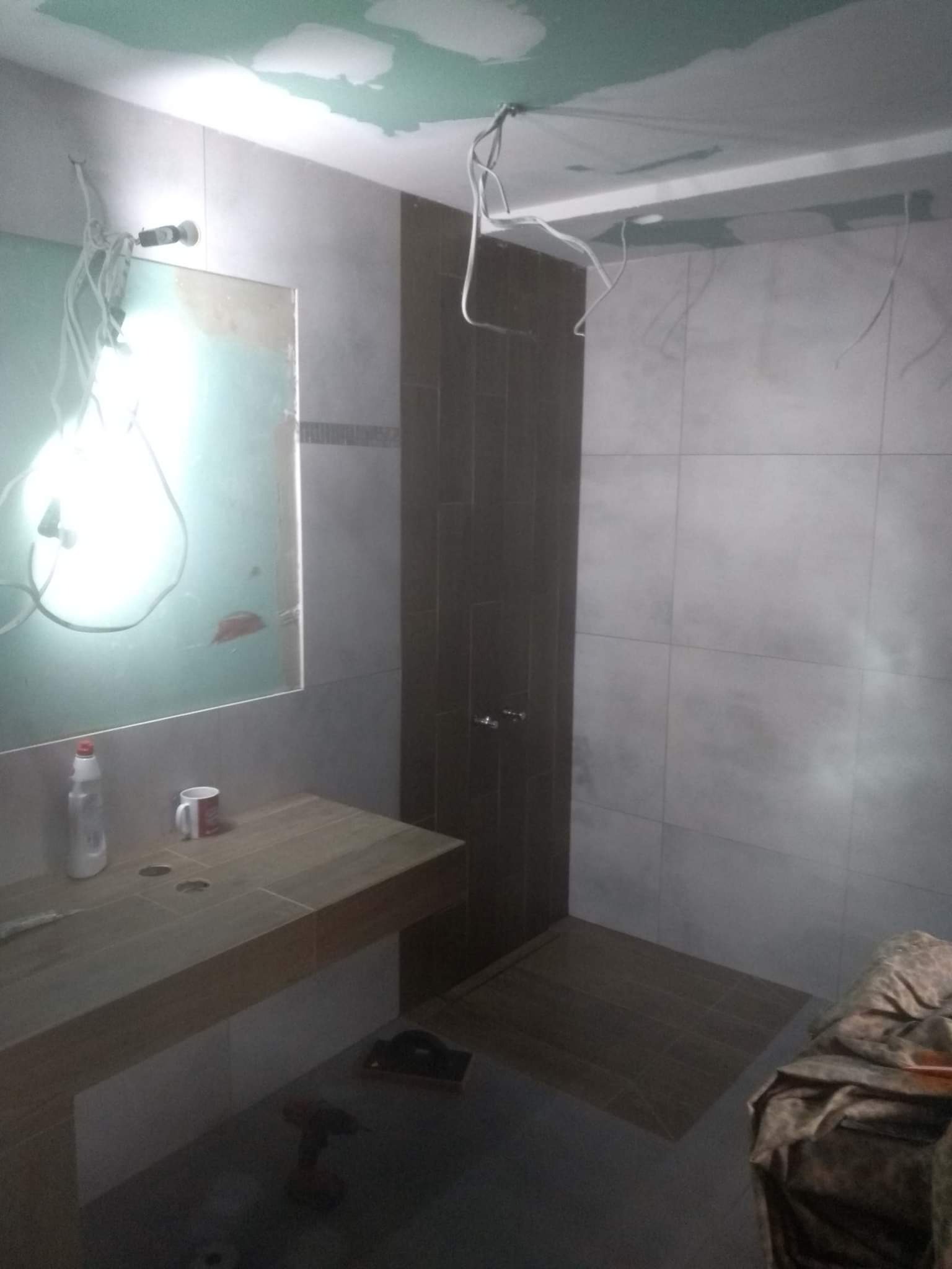 Łazienka w trakcie remontu: widoczne niedokończone płytki, przewody elektryczne, niezabezpieczone lustro, umywalka z drewnianym blatem, narzędzia na podłodze.