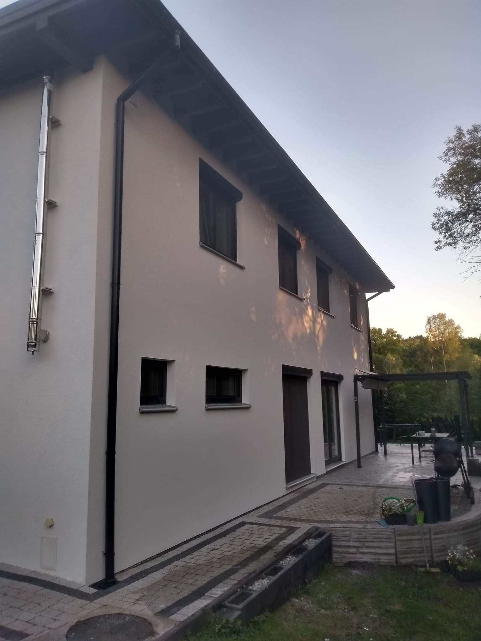 Dwukondygnacyjny dom z jasną elewacją, ciemnymi ramami okiennymi i chromowanym kominem na bocznej ścianie, widok z niskiej perspektywy na taras i zieleń w tle.