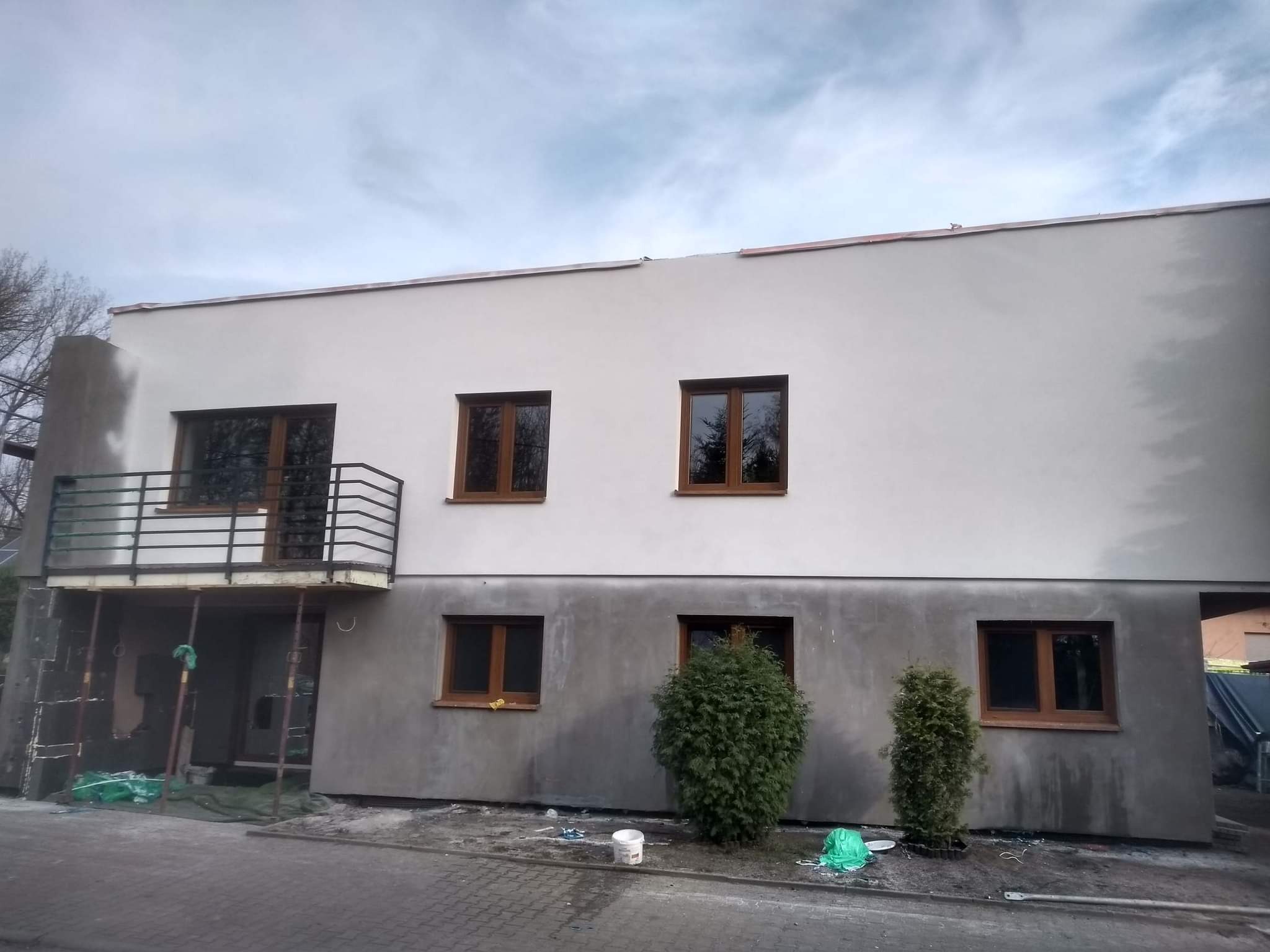 Dwukondygnacyjny budynek z elewacją w trakcie prac: górna część pomalowana na biało, dolna w szarym tynku, widoczne brązowe okna, balkon z czarną balustradą, rusztowanie i dwie małe tuje.