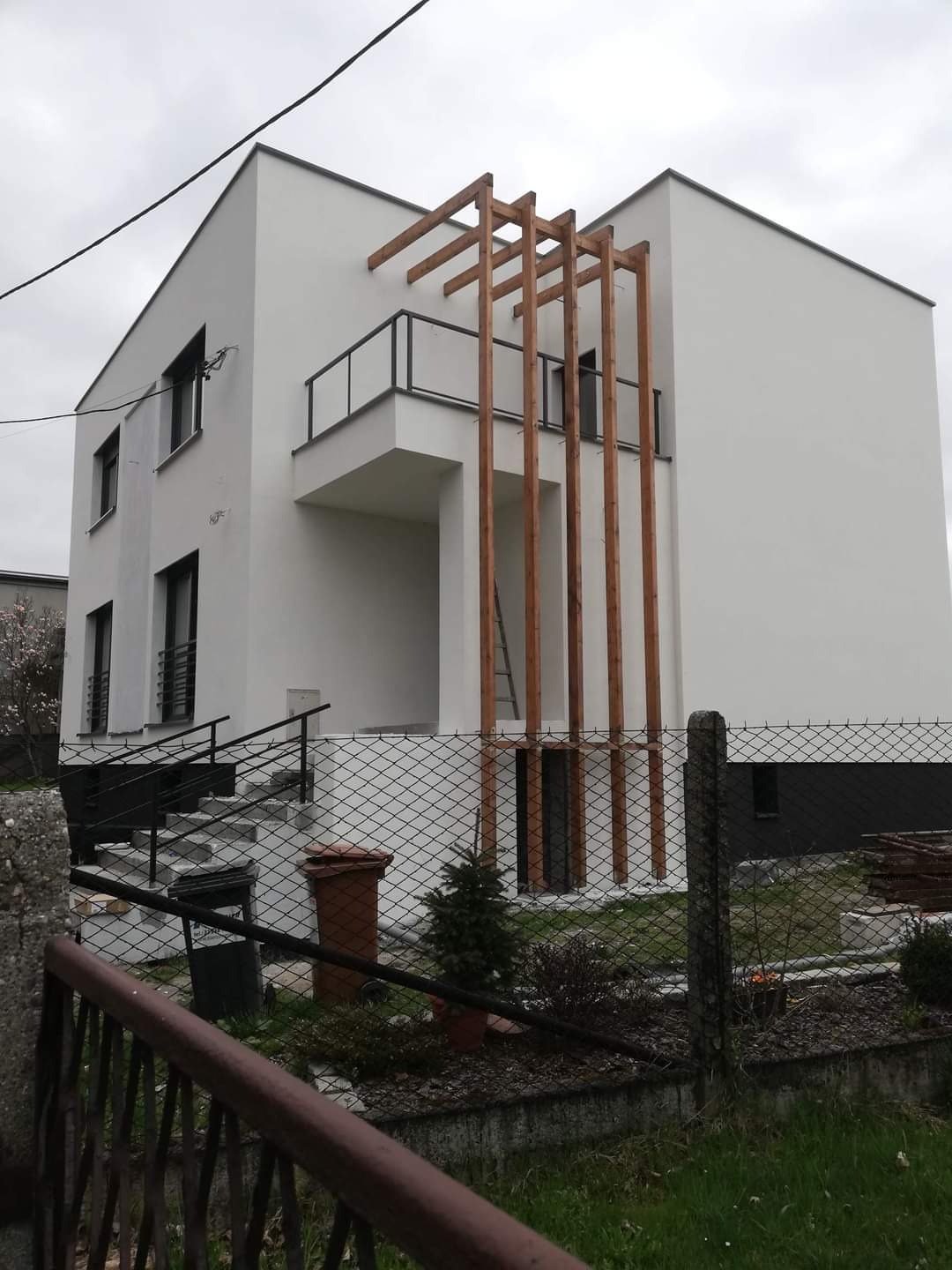 Nowoczesny, biały dom jednorodzinny z drewnianą pergolą nad balkonem, widok z zewnątrz przez siatkę ogrodzeniową.