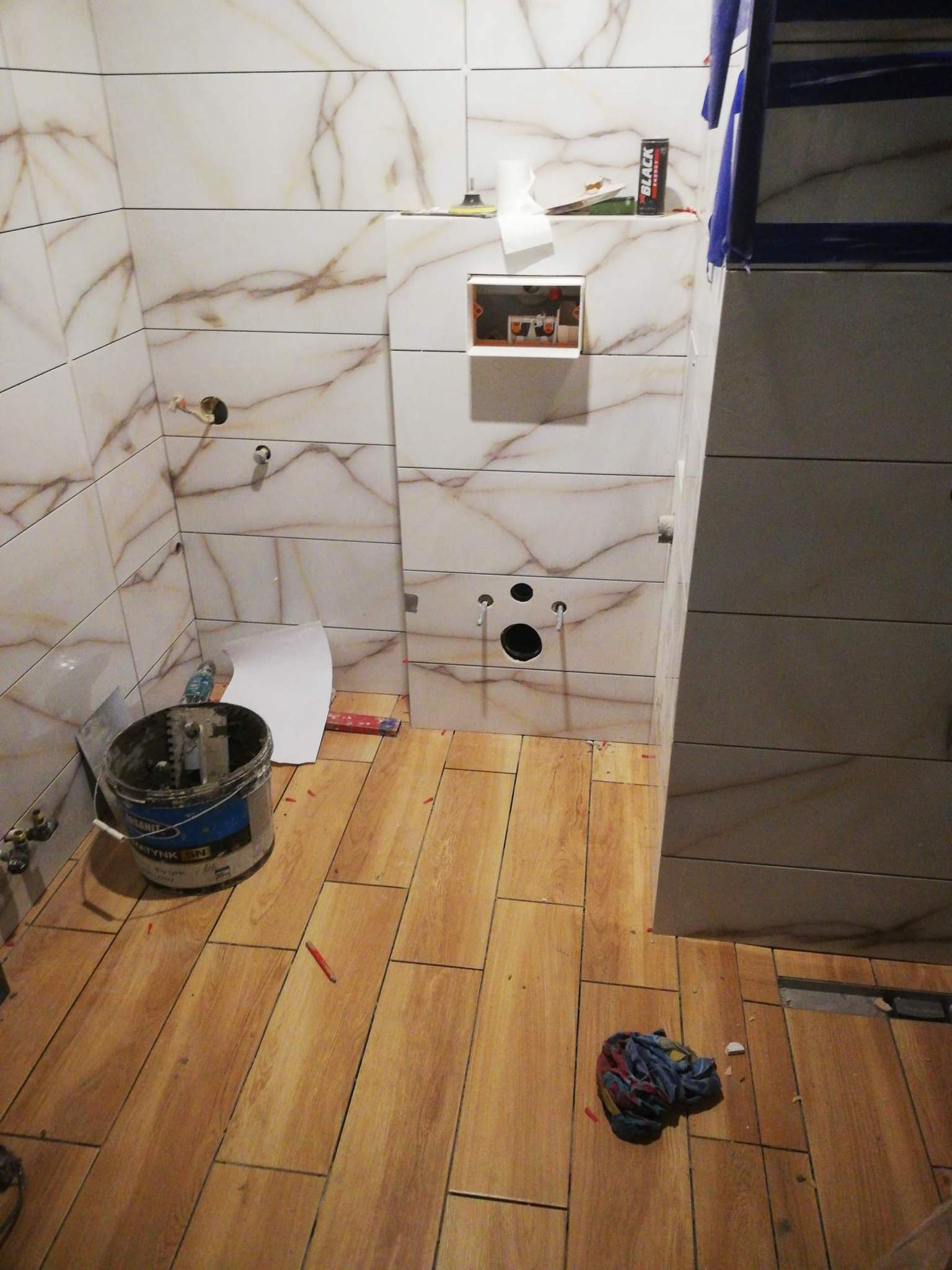 Łazienka w trakcie remontu: białe płytki imitujące marmur na ścianach, drewnopodobne płytki na podłodze, widoczny kubeł z zaprawą, puszka podtynkowa i częściowo zamontowany stelaż podtynkowy WC.