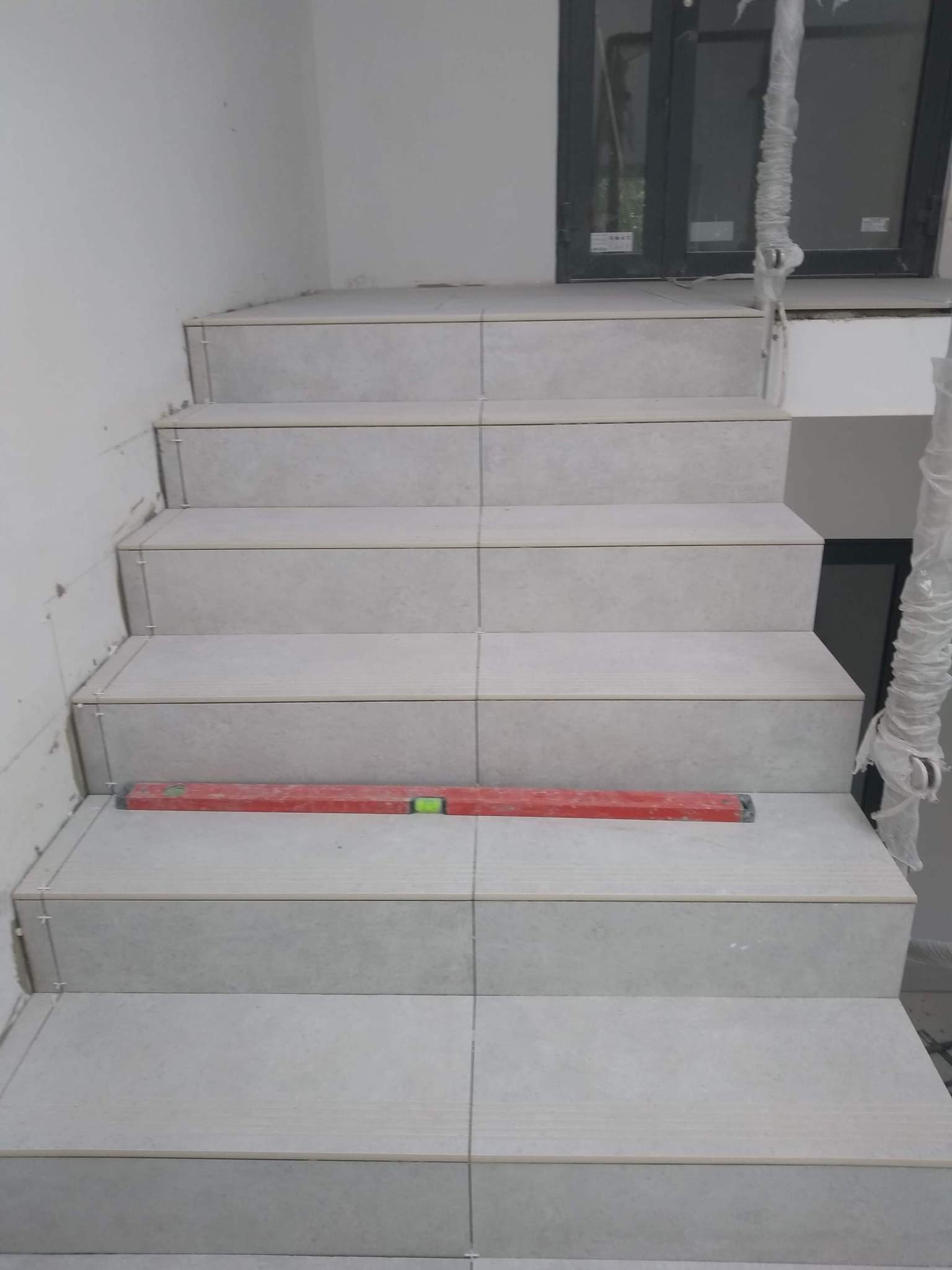 Montaż płytek na schodach wewnętrznych, poziomica murarska na jednym ze stopni, widoczne krzyżyki dystansowe, okno w tle zabezpieczone folią.