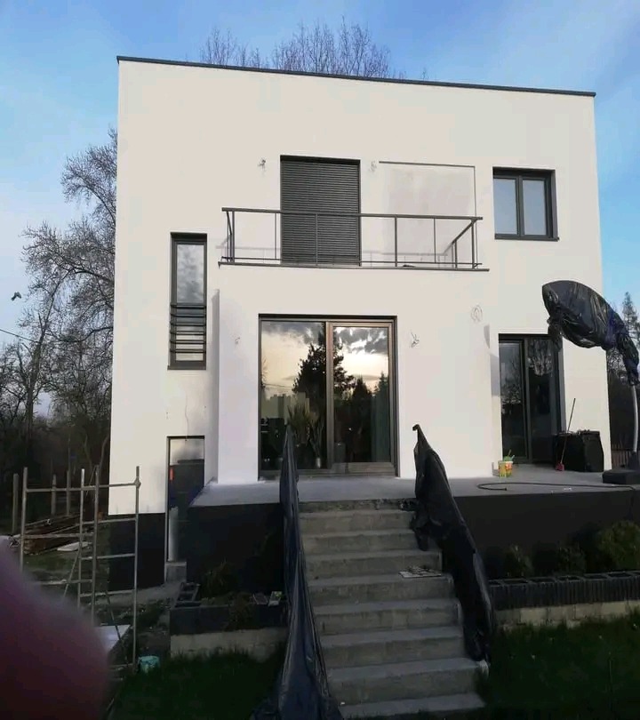 Dwupiętrowy dom z białymi ścianami i szarymi detalami, widoczne schody wejściowe zabezpieczone folią malarską, ślady świeżego malowania na elewacji i podłożu, drabina oparta o ścianę z lewej strony.