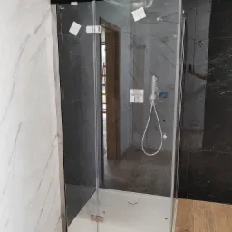 Nowoczesna kabina prysznicowa z przezroczystego szkła, zamontowana w łazience z marmurowymi płytkami w odcieniach bieli i szarości, widoczne chromowane zawiasy i biały brodzik, elementy instalacji...