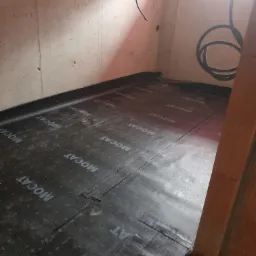 Przygotowanie podłoża pod posadzkę: czarna mata z nadrukowanym wzorem kratki i logo MOCAT, rozłożona na betonowej podłodze w rogu pomieszczenia, widoczne kable elektryczne na ścianie.