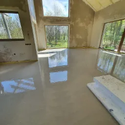 Świeżo wylana, mokra posadzka jastrychowa w surowym wnętrzu domu jednorodzinnego z widokiem na ogród przez duże okna; widoczne płyty styropianowe.