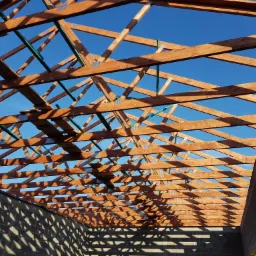 Konstrukcja dachu z drewnianych belek, połączonych metalowymi płytkami, widoczna od wewnątrz budynku w stanie surowym, na tle błękitnego nieba.