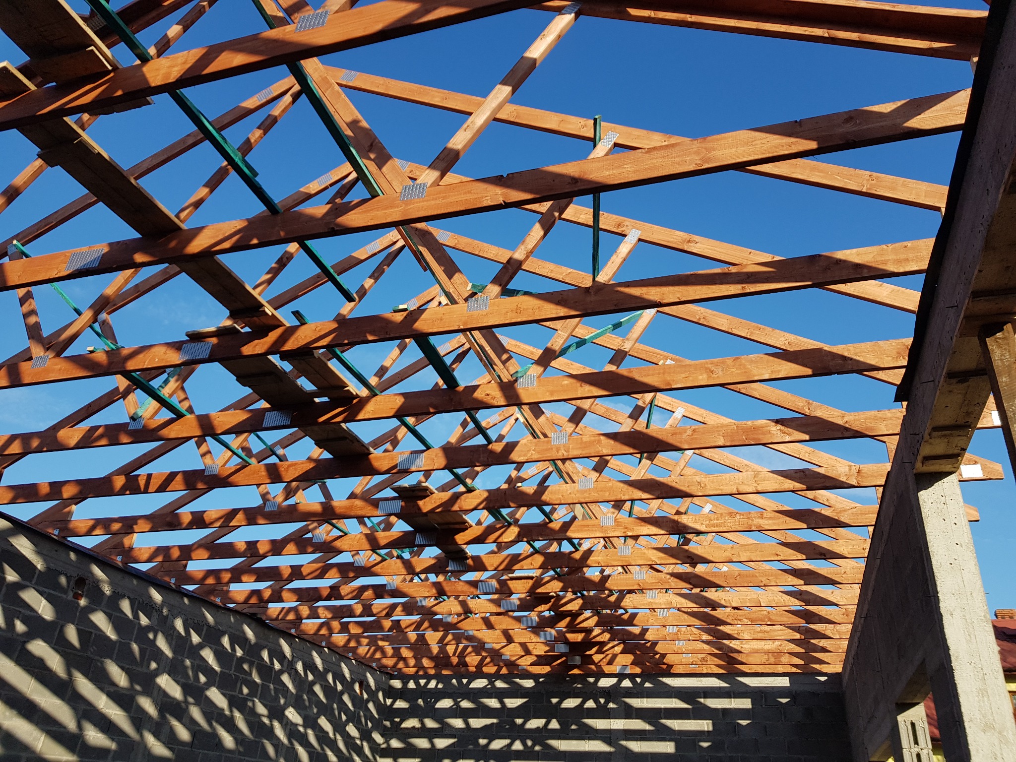 Konstrukcja dachu z drewnianych belek, połączonych metalowymi płytkami, widoczna od wewnątrz budynku w stanie surowym, na tle błękitnego nieba.