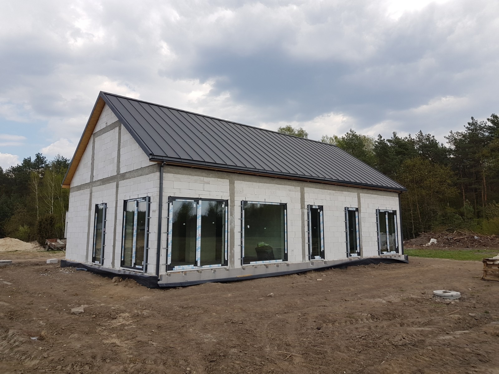 Surowy stan domu w trakcie budowy: ściany z białych bloczków, czarny dach z blachy trapezowej, okna z folią ochronną na tle pochmurnego nieba.