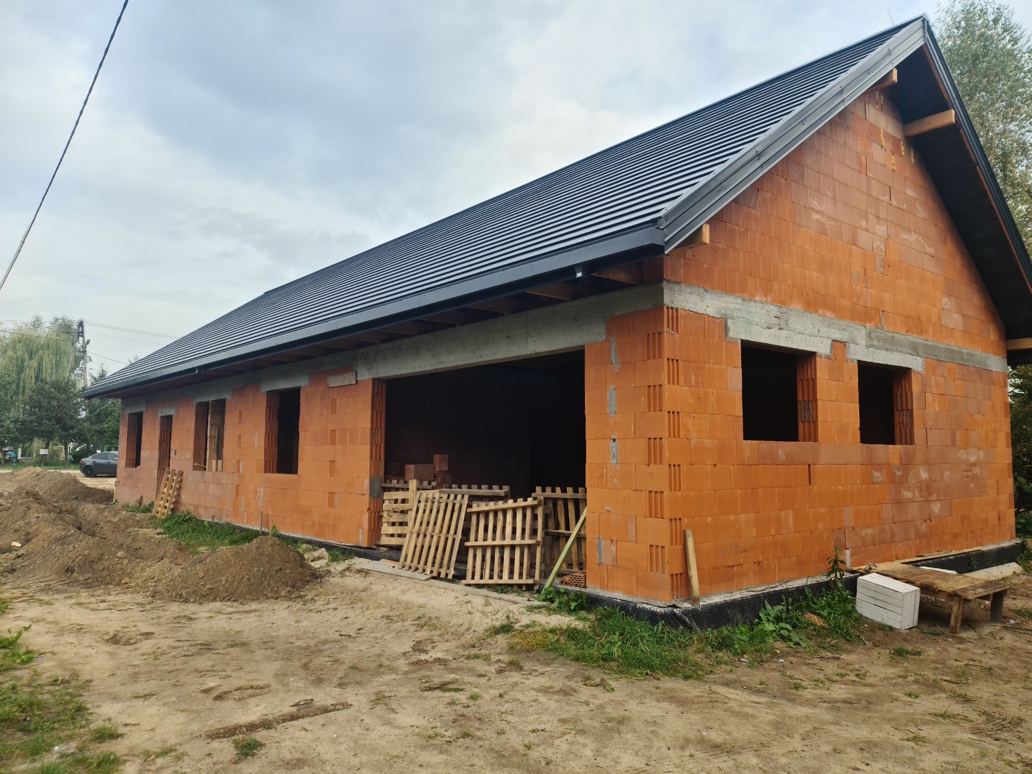 Niedokończony budynek z czerwonej cegły z czarnym dachem z blachy na etapie budowy, widoczne palety i pustaki wewnątrz.