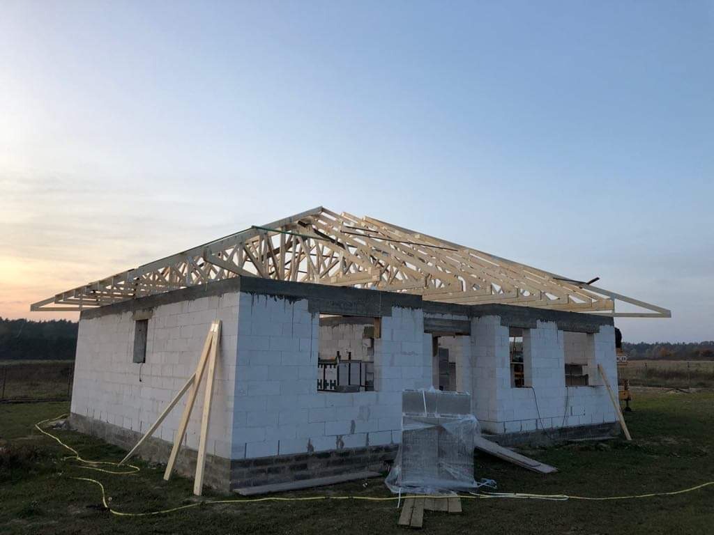 Surowy budynek z białego bloczka z drewnianą konstrukcją dachu w trakcie montażu, widoczne elementy wiązarów, w tle pole i las przy zachodzącym słońcu.