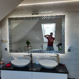 Nowoczesna łazienka na poddaszu z dwoma umywalkami nablatowymi, lustrem z podświetleniem LED i ramą w szachownicę, oraz widocznym odbiciem fotografa robiącego zdjęcie.