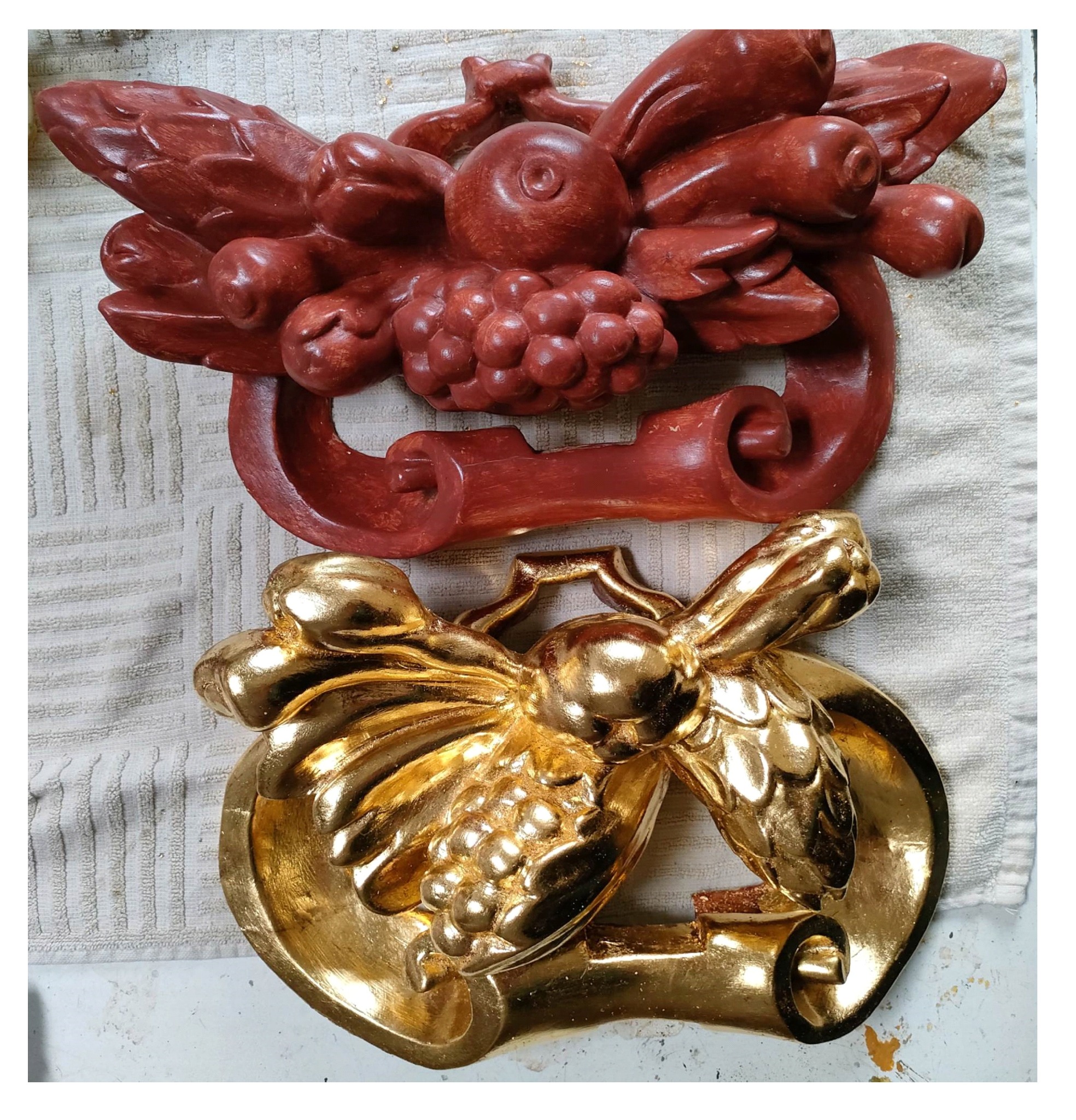 Dwie drewniane płaskorzeźby z motywem owoców, jedna pomalowana na czerwono-brązowy kolor, druga pozłocona, leżą na tekstylnej powierzchni.