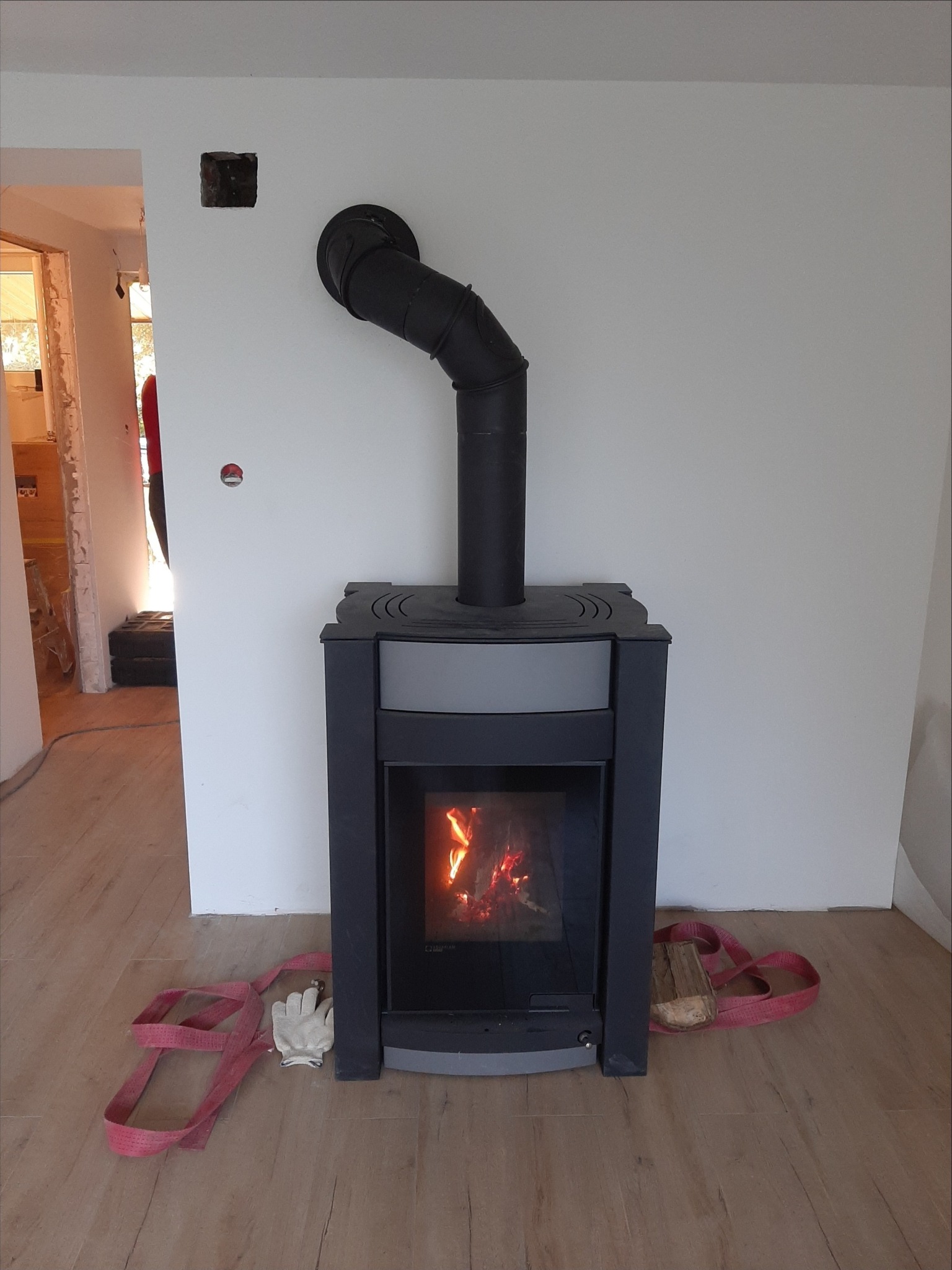 Nowoczesny piec wolnostojący z czarnym przewodem kominowym, płomienie widoczne przez szybę, rękawice robocze i pas transportowy leżące na podłodze z paneli.