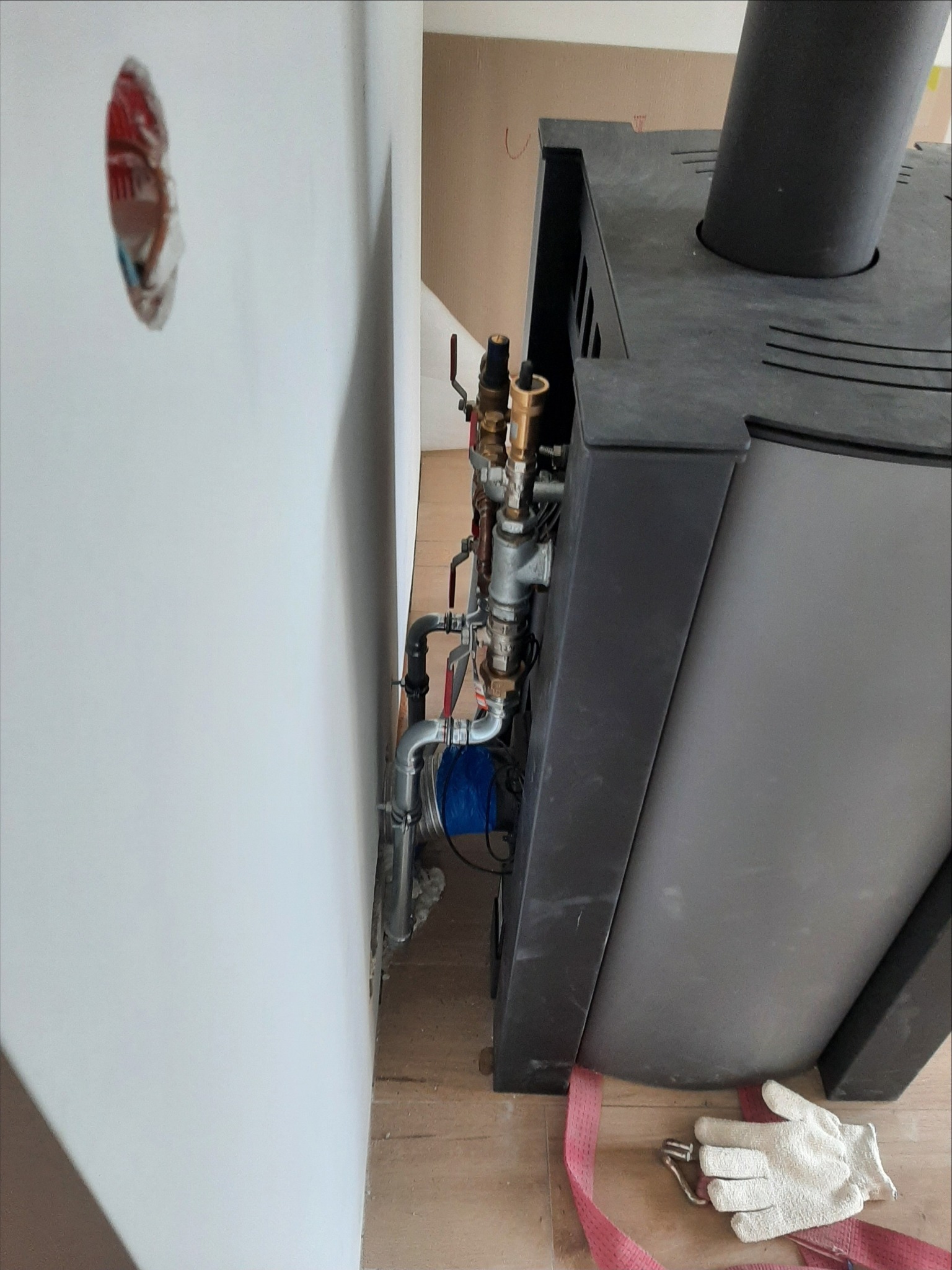 Szara obudowa pieca z podłączonymi rurami i zaworami, widoczne białe rękawice robocze i różowy pas transportowy na drewnianej podłodze, ściana z widocznymi przewodami.