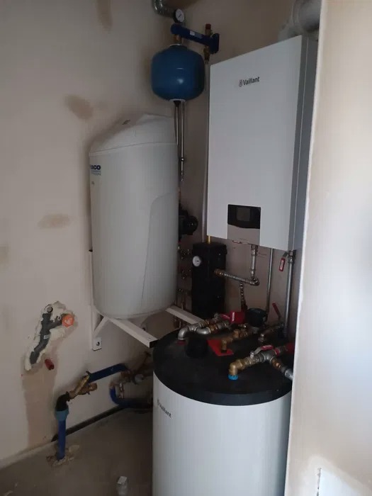 Instalacja grzewcza w kotłowni: biały zasobnik ciepłej wody z czarnym blatem, wiszący biały piec gazowy Vaillant, niebieskie naczynie wzbiorcze, rury miedziane i plastikowe, ściany w trakcie...