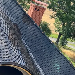 Czarny dach z łuski z kominem z cegły na tle polnego krajobrazu, widoczne miedziane elementy mocujące.