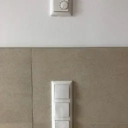 Biały panel z trzema włącznikami światła na tle beżowych płytek oraz regulator temperatury na białej ścianie powyżej.