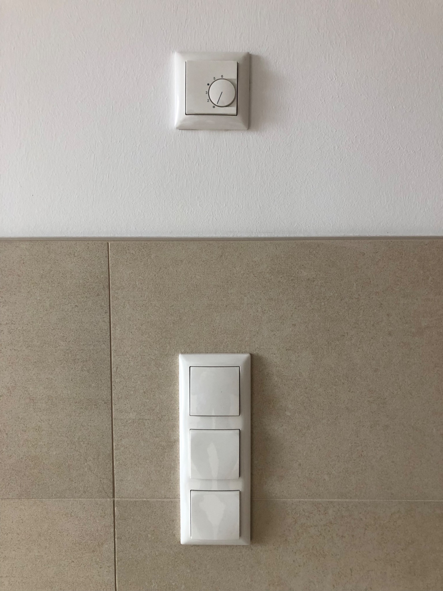 Biały panel z trzema włącznikami światła na tle beżowych płytek oraz regulator temperatury na białej ścianie powyżej.