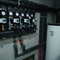 INSTAL AQUA Waldemar Dzierżak - Rozdzielacz ogrzewania podłogowego z manometrami, pompami obiegowymi i zaworami, oraz biały zasobnik ciepłej wody Kospel, rury w izolacji na szarej ścianie.