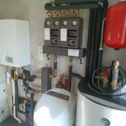 Kompletna kotłownia z piecem gazowym, zasobnikiem, grupą pompową Warmit i filtrem. Widoczne rury, zawory i manometry. Nowoczesna instalacja grzewcza w pomieszczeniu.