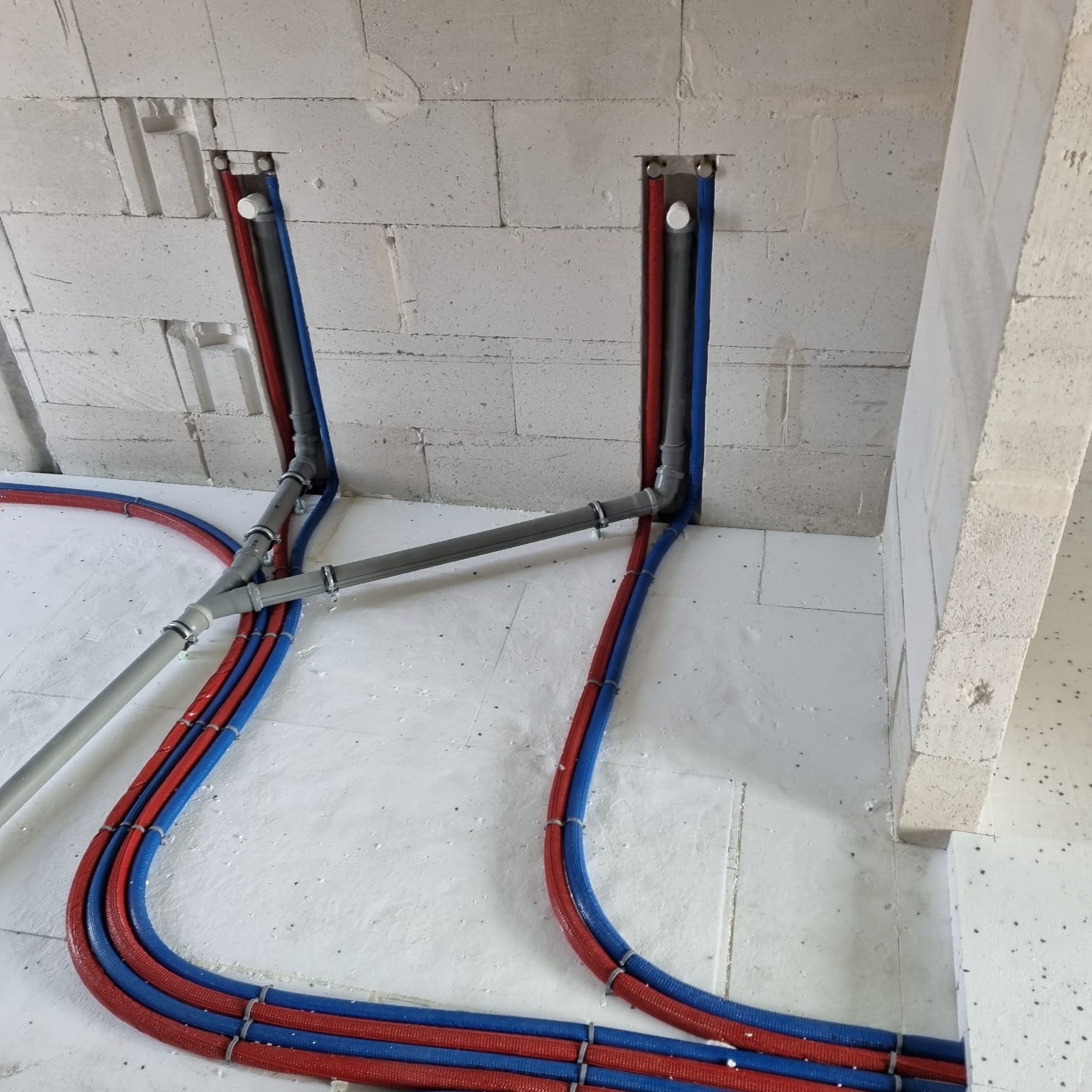 Instalacja rur grzewczych (czerwone i niebieskie) oraz szarych rur kanalizacyjnych w bruzdach w ścianie z betonu komórkowego i na podłodze z izolacją.