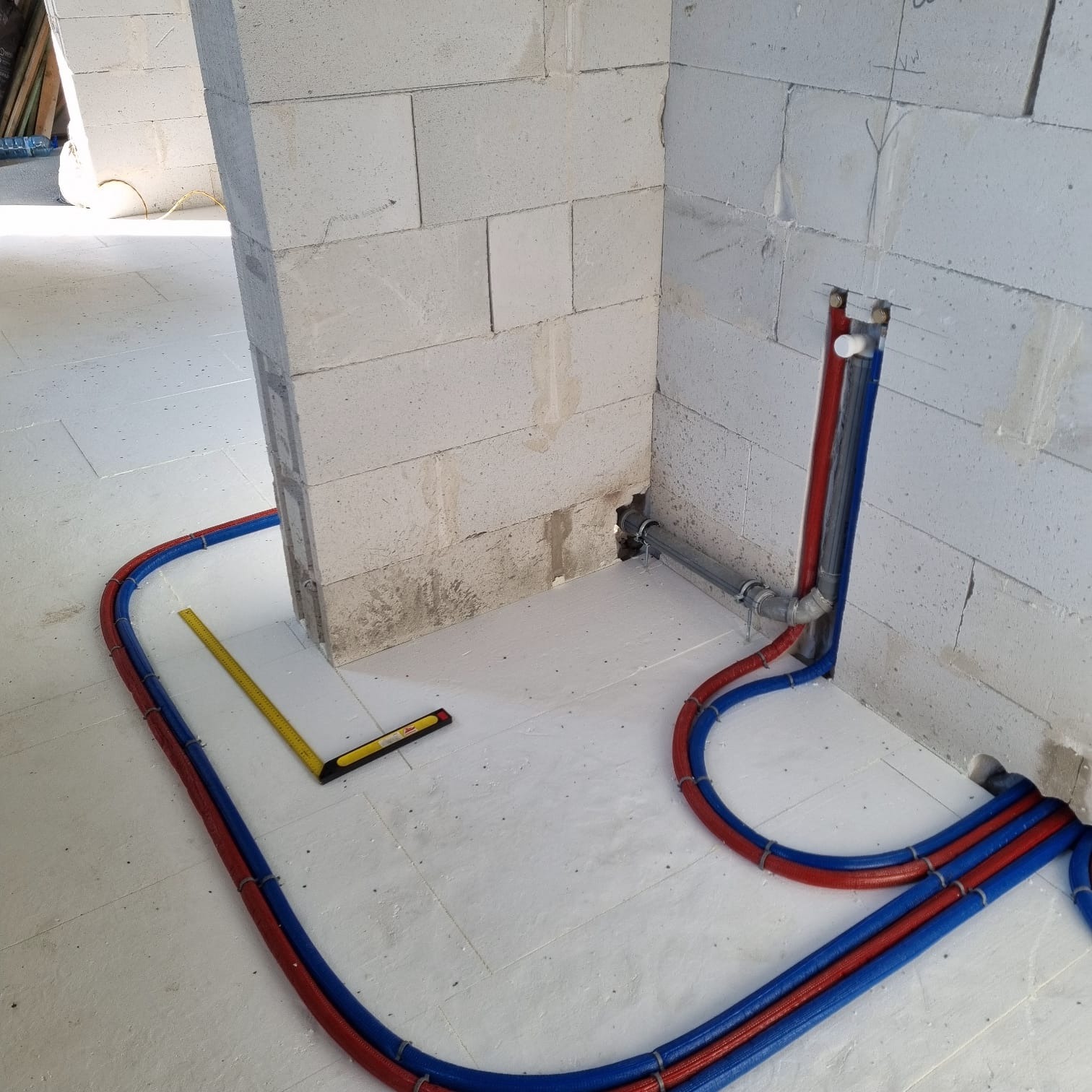 Instalacja rur (czerwone, niebieskie) na białej izolacji podłogowej w surowym wnętrzu. Widoczny poziomica. Przygotowanie do wylewki.