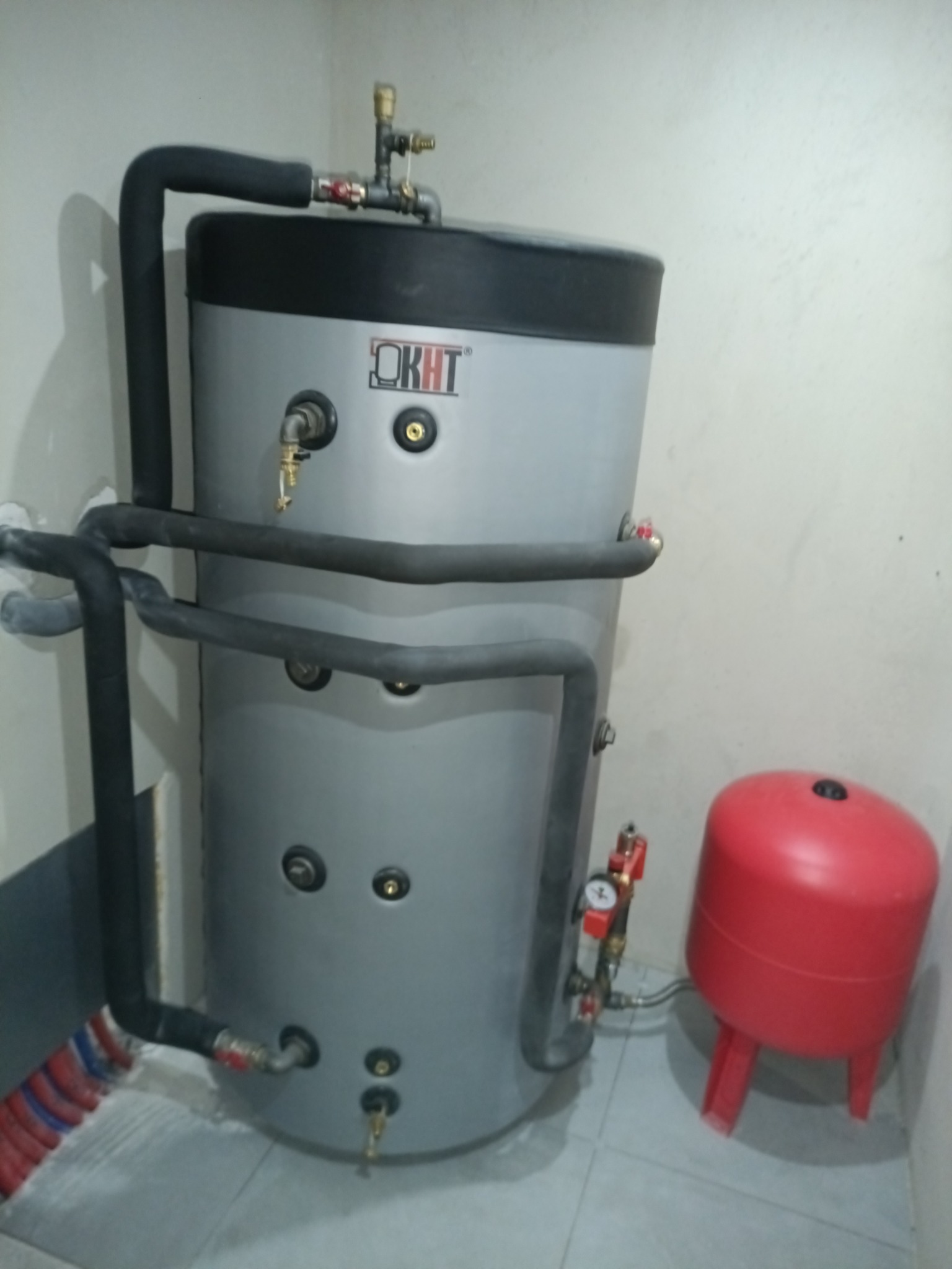 Szary zasobnik ciepłej wody użytkowej OKHT z czarnymi rurami, manometrem i czerwonym naczyniem przeponowym w pomieszczeniu z jasnymi ścianami i szarą podłogą.