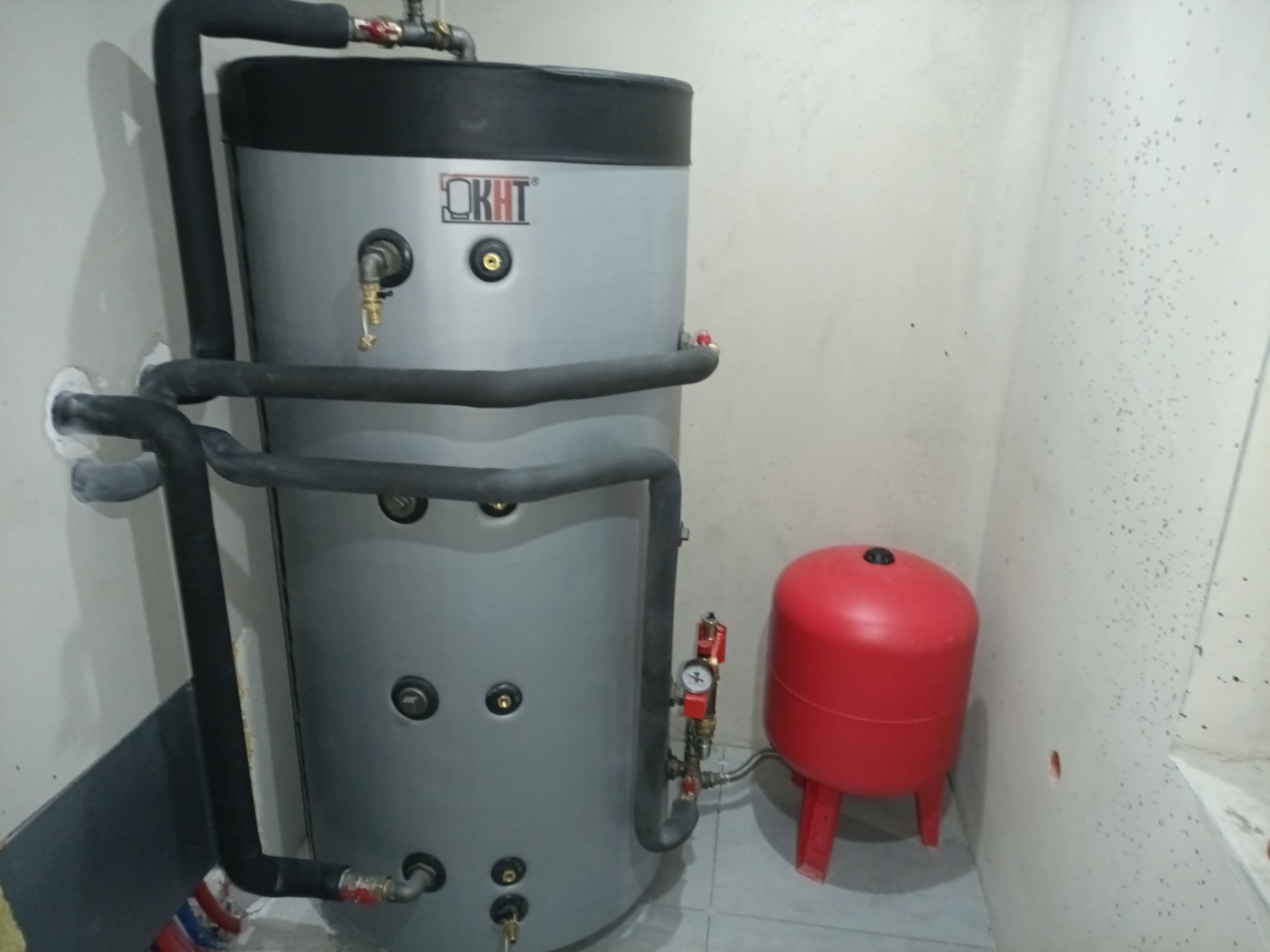 Szary bojler KHT z czarnym ociepleniem i czerwony zbiornik wyrównawczy w rogu pomieszczenia. Instalacja hydrauliczna z zaworami i manometrem.