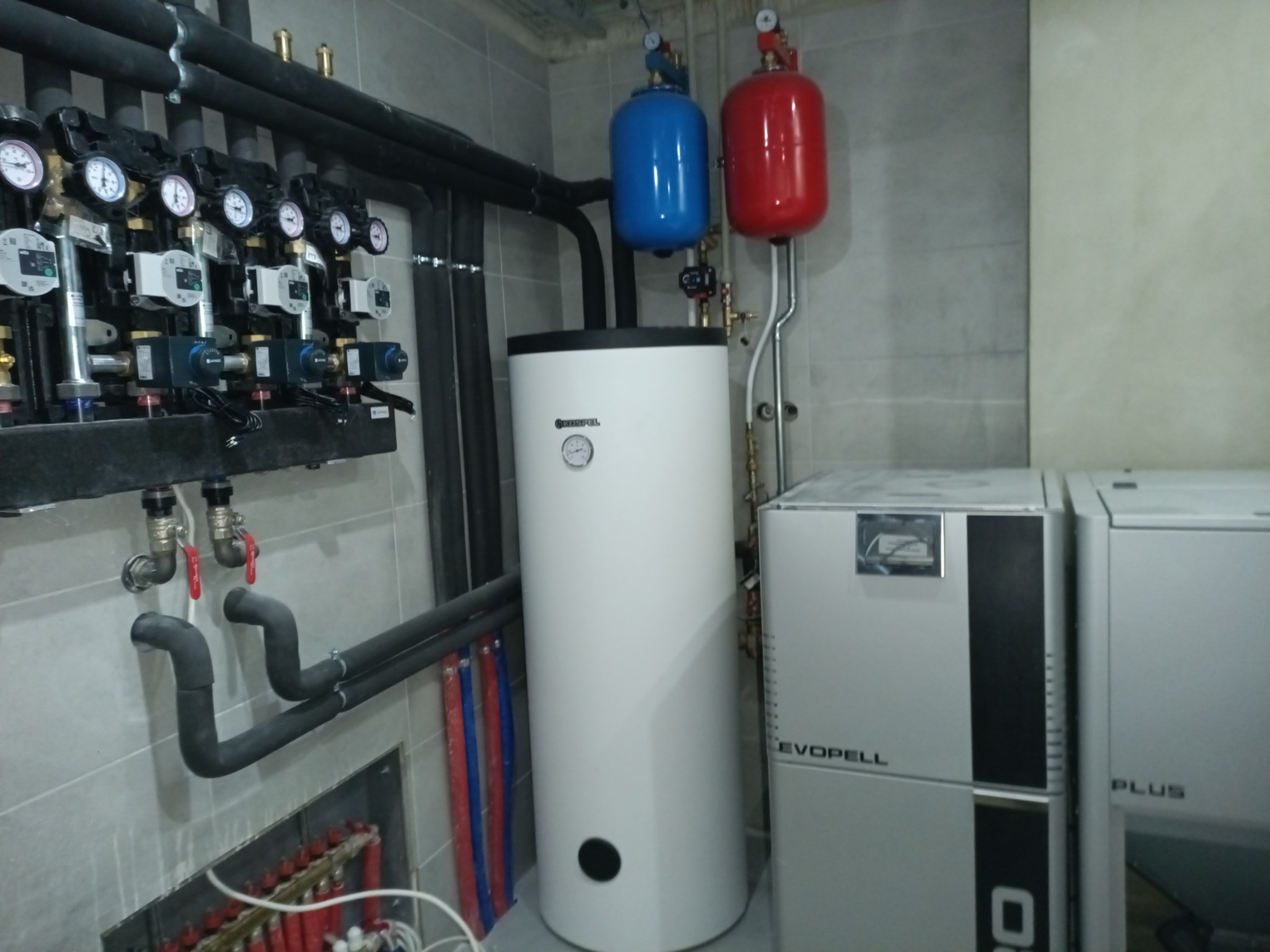 Biały zasobnik ciepłej wody użytkowej Kospel, z czarnym otworem rewizyjnym, na tle szarej ściany z instalacją CO, z czerwonym i niebieskim zbiornikiem wyrównawczym.