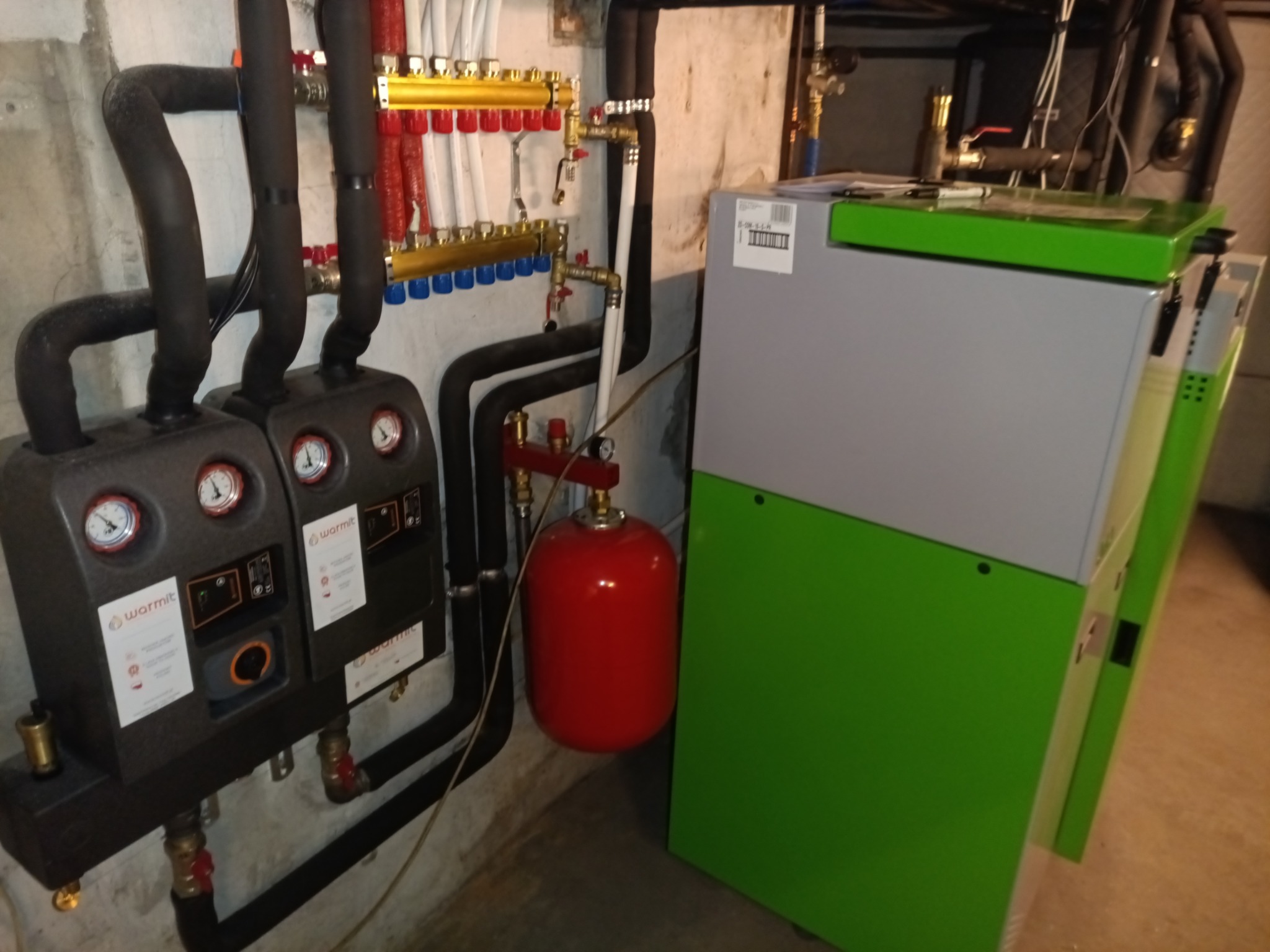 Nowoczesny piec z zielonymi akcentami, podłączony do instalacji grzewczej z rozdzielaczem i czarnymi rurami, oraz czerwonym zbiornikiem wyrównawczym.