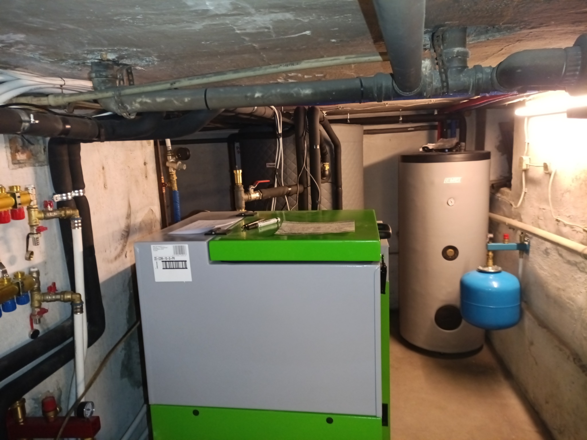 Nowoczesny piec CO z zielonym akcentem i zasobnikiem ciepłej wody w kotłowni. Widoczne rury instalacji i osprzęt.