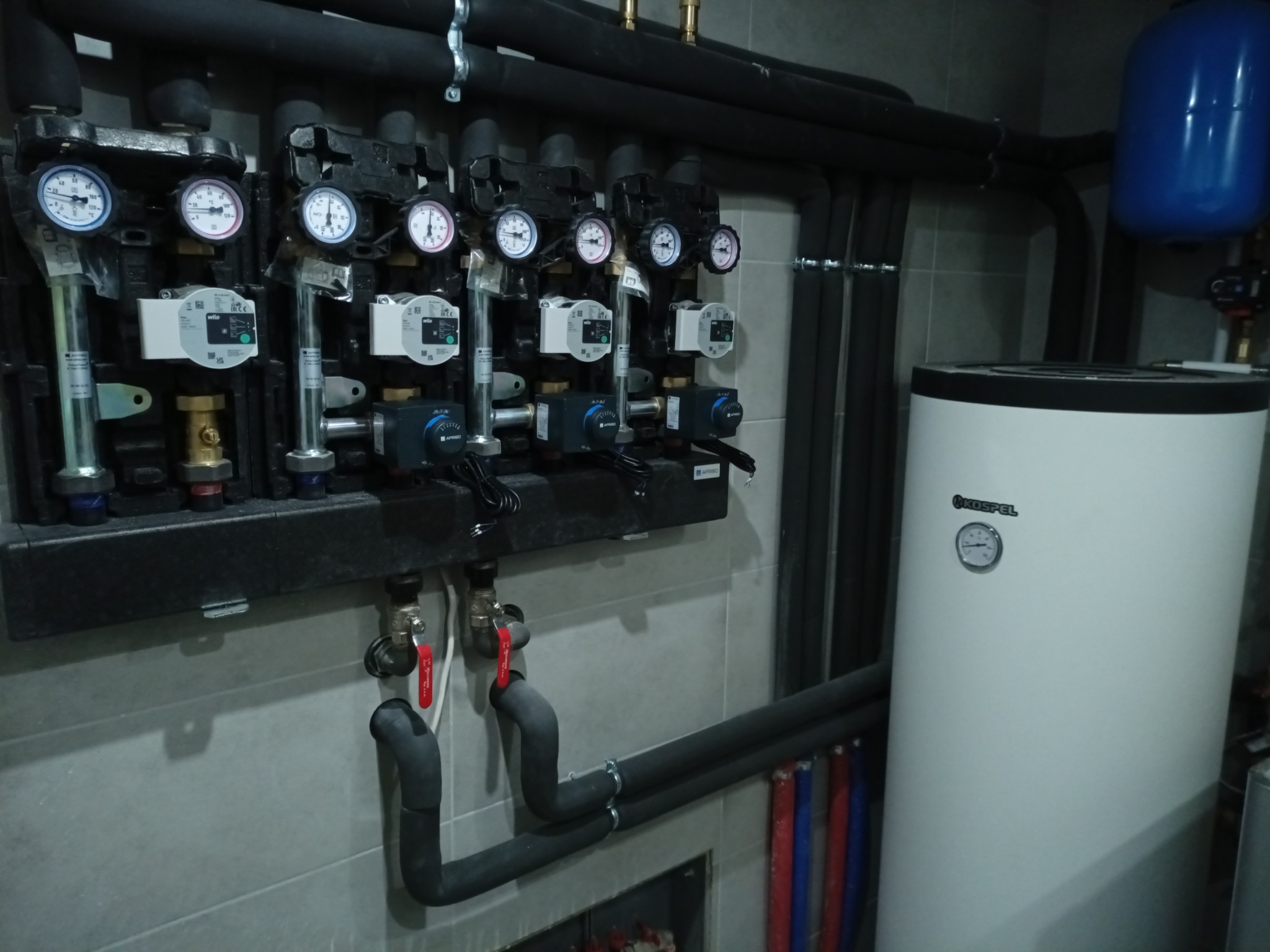 Rozdzielacz ogrzewania podłogowego z manometrami, pompami obiegowymi i zaworami, oraz biały zasobnik ciepłej wody Kospel, rury w izolacji na szarej ścianie.