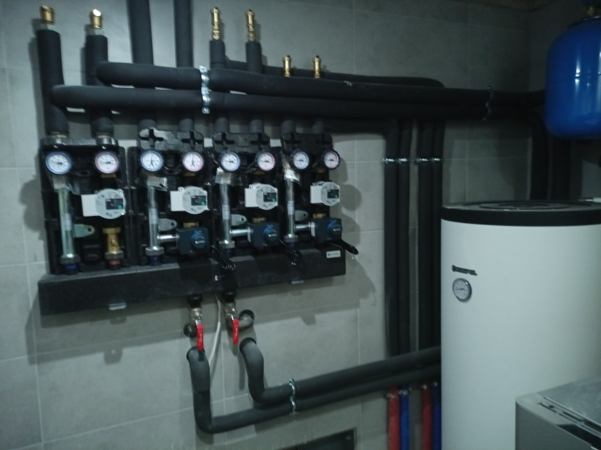 Rozdzielacz ogrzewania podłogowego z manometrami i rurami w Bytomiu. Widoczny również zasobnik ciepłej wody i niebieski zbiornik wyrównawczy.