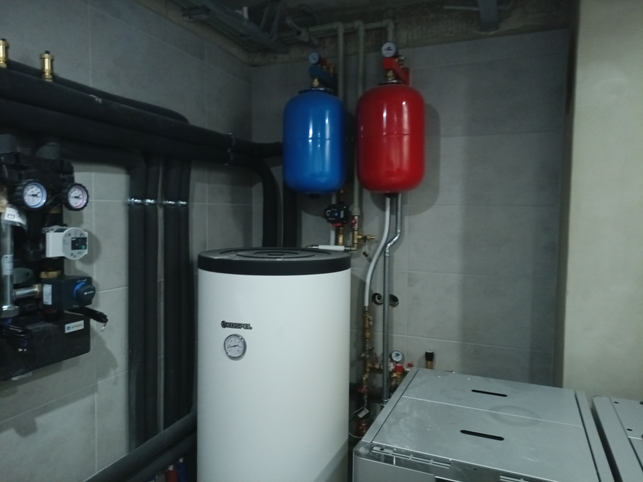 Bojler Kospel z instalacją: czerwony i niebieski zbiornik wyrównawczy ciśnienia, rury w izolacji, manometry. Całość na tle szarej ściany z płytek.