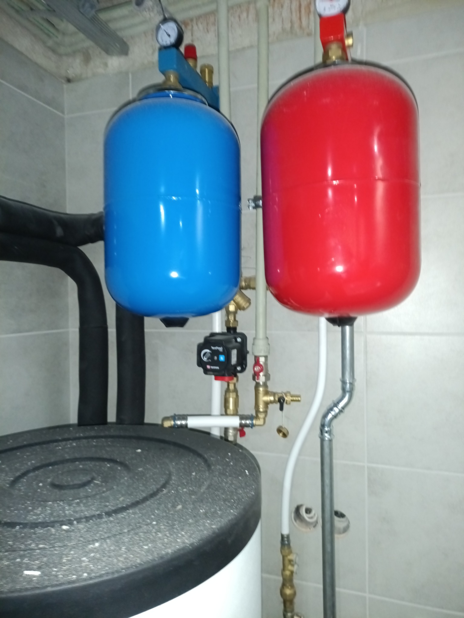 Instalacja CO z czerwonym i niebieskim zbiornikiem wyrównawczym, widoczne rury, zawory i manometry. Montaż w kotłowni, płytki na ścianie.